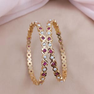 Może przedstawiać: Dwie złote bransoletki typu bangle z naprzemiennym wzorem okrągłych czerwonych rubin&oacute;w i białych pereł osadzonych w geometrycznej, fasetowanej metalowej ramie. Okrągłe bransoletki mają polerowane wykończenie i są ustawione pionowo.