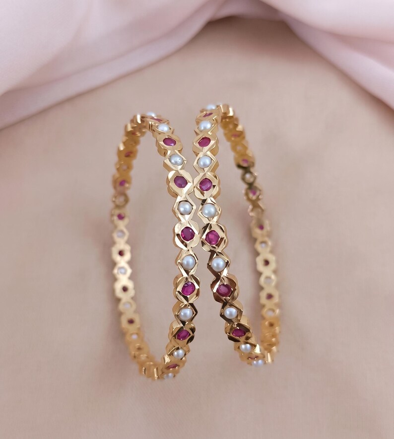 Może przedstawiać: Dwie złote bransoletki typu bangle z naprzemiennym wzorem okrągłych czerwonych rubin&oacute;w i białych pereł osadzonych w geometrycznej, fasetowanej metalowej ramie. Okrągłe bransoletki mają polerowane wykończenie i są ustawione pionowo.