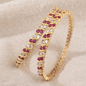 Può includere: Due bracciali rigidi dorati caratterizzati da un motivo ripetitivo di pietre ovali rosso scuro alternate a pietre rotonde trasparenti incastonate in un design in metallo intrecciato. I bracciali hanno una finitura lucida e sono disposti.
