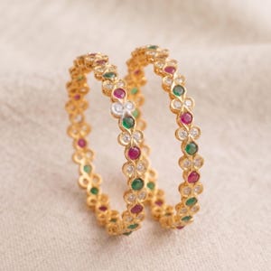 May include: Two gold-toned bangles featuring a repeating floral-inspired pattern. Each link is set with alternating round-cut red, green, and clear white stones. The metal structure creates a delicate, scalloped edge along the circular band.