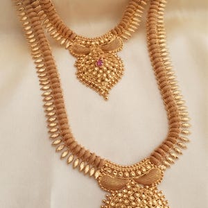 May include: Two gold-toned necklaces featuring a repeating leaf-like pattern along the chains. Each necklace terminates in a large, ornate pendant with a central circular or heart-shaped design, embellished with a single small, round, deep pink stone.
