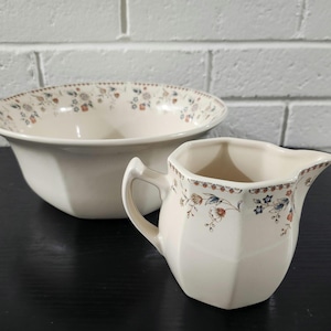 May include: A cream-colored ceramic bowl and a matching creamer pitcher, both featuring a delicate floral pattern in shades of blue, orange, and brown along the upper rims. The pitcher has an angular handle and a pointed pouring spout.