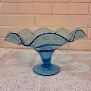 May include: A translucent blue glass compote dish featuring a wide, ruffled, scalloped rim and a smooth, circular pedestal base. The bowl transitions from a lighter, airy blue at the top edge to a deeper, more saturated blue at the base.