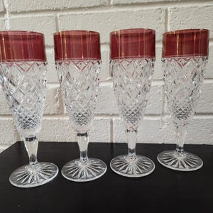 May include: A set of four clear crystal champagne flutes featuring a diamond-cut pattern on the lower bowls and stems, topped with a smooth, deep red glass rim. Each glass stands on a circular base with a radiating starburst design.