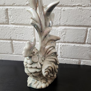 May include: A ceramic rooster figurine featuring an iridescent white glaze with gold-painted trim on the feathers, comb, and tail. The rooster stands in an upright position with a prominent, stylized tail extending upward.