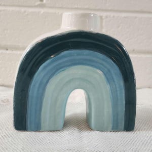 May include: A ceramic vase shaped like an arch with a glossy finish. The surface features concentric bands of color in shades of dark navy blue, medium blue, and light pale blue, with a solid white top section surrounding the circular opening.