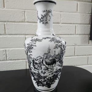 May include: A white ceramic vase featuring a black toile-style print of a rooster, hens, and chicks in a rural landscape. The vase has a slender neck, a flared rim with a black edge, and a rounded body decorated with detailed floral and farm motifs.