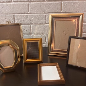 May include: A collection of five picture frames of varying shapes and sizes. The set includes a gold octagonal frame, a tall rectangular gold frame, a small gold frame, a large gold frame with a dark border, and a dark brown rectangular frame.