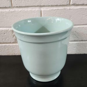 May include: A pale mint green ceramic planter with a smooth, glossy finish. The vessel features a wide, open circular rim that tapers down to a narrow, raised circular base, creating a classic, rounded silhouette with a subtle horizontal ridge.