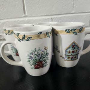 May include: A set of three white ceramic mugs featuring a decorative border of green leaves on a tan band. Each mug displays a different garden-themed illustration, including a birdhouse with hearts, a potted plant with a bee, and floral motifs.