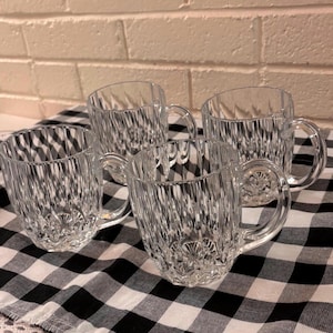 May include: A set of four clear glass mugs featuring a vertical ribbed texture on the exterior walls and a starburst pattern on the base. Each mug has a curved handle and a wide, circular rim, showcasing a transparent design throughout the set.