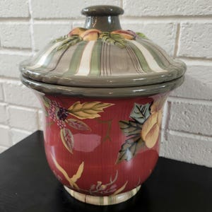 May include: A ceramic canister with a lid featuring a red base decorated with painted fruit and foliage. The lid has a striped pattern in shades of green, white, and brown with a sculpted fruit finial. The canister has a wide rim and a base.