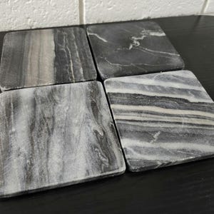 May include: A set of four square coasters made of natural stone, each featuring a unique pattern of black, gray, and white veining. The coasters have smooth, flat surfaces and rounded edges, displaying distinct marble-like textures and color variations.