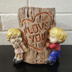 May include: A brown tree stump planter featuring a carved heart with the text I LOVE YOU. Two blonde children are attached to the sides, one wearing a pink dress and the other wearing a red shirt with blue pants, both with painted facial features.