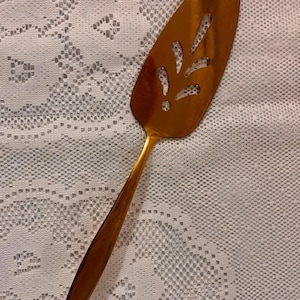 May include: A gold-toned metal cake server featuring a triangular blade with a pointed tip and a long, slender handle. The blade is decorated with a series of cut-out leaf-shaped patterns arranged in a symmetrical design along the center.