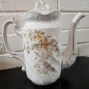 May include: A white porcelain teapot featuring a delicate floral pattern in shades of yellow, brown, and green. The teapot has a swirled, ribbed body, a scalloped rim, an ornate handle, a curved spout, and a matching lid with a decorative finial.