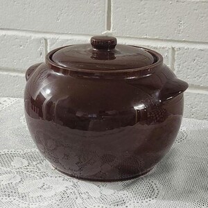 May include: A round, dark brown ceramic pot with a glossy finish, featuring a matching lid with a small central knob and two integrated side handles. The pot has a smooth, curved body and a deep, uniform chocolate-colored glaze throughout.