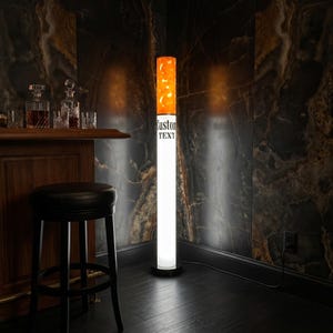 May include: A tall, cylindrical floor lamp shaped like a cigarette, featuring a glowing orange top section and a white lower section with the black text "Custom TEXT" printed on the side, standing on a circular black base in a dark room.