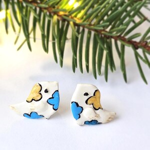 May include: A pair of white stud earrings shaped like small birds. Each bird features hand-painted details including a gold-colored head patch, a bright blue wing section, and a small black eye. The earrings have a smooth, matte finish.