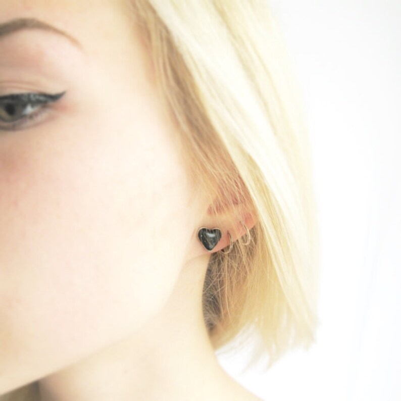 May include: A close-up of a person's ear with a small black heart-shaped earring and a silver hoop earring.