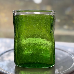 Può includere: Un vaso quadrato di vetro verde con una superficie strutturata. Il vaso è riempito con un liquido verde e poggia su un piatto grigio e circolare. Il vetro ha una consistenza a bolle e la luce lo attraversa.