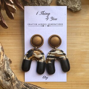 May include: A pair of handmade earrings with a unique design. The earrings feature a circular top in a bronze color, connected to a black and white arch shape. The text "1 Thing 4 You" and "Crafted Across Generations" is visible on the card.