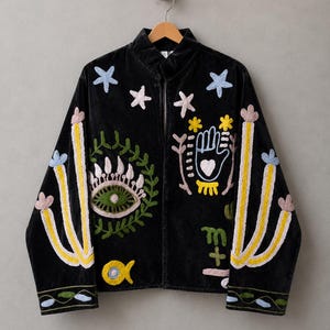 May include: A black velvet jacket featuring colorful embroidered motifs including stars, a green eye, a hand with a heart, a yellow fish, and candelabra designs on the sleeves. The jacket has a mandarin collar and long sleeves with floral trim.