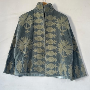 May include: A slate blue velvet jacket featuring light green embroidered floral and abstract patterns. The garment has a high collar, long sleeves, and a front opening. The interior lining is visible at the bottom hem, showing a white and blue striped pattern.