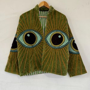 May include: An olive green jacket featuring large embroidered eye motifs on the front and sleeves, with orange radiating lines. The jacket has a stand-up collar and an interior lining with a vertical light green and white striped pattern.