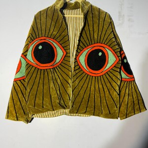 May include: An olive green velvet jacket featuring large embroidered eye motifs on the front and sleeves. The eyes have black pupils, light green irises, and orange outlines, with black radiating lines. The jacket has a vertical striped lining.