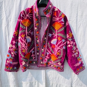 Pode incluir: Um casaco de veludo magenta com bordados florais multicoloridos intrincados em tons de laranja, rosa, azul claro e verde. O casaco tem gola alta, mangas compridas e uma frente aberta que revela um forro às riscas verticais brancas e azuis.