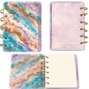 May include: A small ring-bound notebook featuring a marbled cover in shades of purple, blue, and pink with embedded gold-colored flakes. The notebook includes a translucent purple back cover with horizontal lines and white lined paper pages inside.