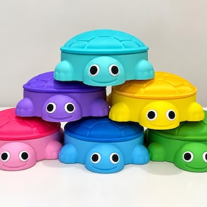 May include: A stack of six turtle-shaped containers in bright colours including turquoise, purple, yellow, pink, blue, and green. Each container features a moulded shell pattern on the lid and a smiling face with large black and white eyes.