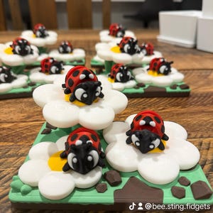 May include: A collection of 3D printed fidget toys featuring red and black ladybirds resting on white flowers with yellow centres, set on green square bases with brown accents. The text @bee.sting.fidgets is printed in white on the bottom right corner.