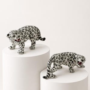 Peut inclure: Deux figurines de léopard en céramique avec une base blanche et des taches noires. Chaque léopard est représenté dans une posture de rôdeur, la gueule ouverte révélant des détails rouges, des dents acérées et une longue queue.