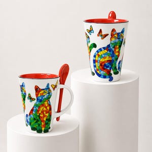 Peut inclure: Deux tasses en céramique blanche ornées d'un motif de chat et de papillons style mosaïque colorée. Chaque tasse possède un intérieur rouge vif et une cuillère rouge assortie qui s'insère dans une fente sur l'anse de la tasse.