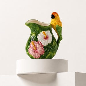 May include: A ceramic jug featuring a green leaf-textured surface adorned with two hibiscus flowers in white and pink. The handle is shaped like a yellow and green parrot with an orange head and a black beak, integrated into the jug design.