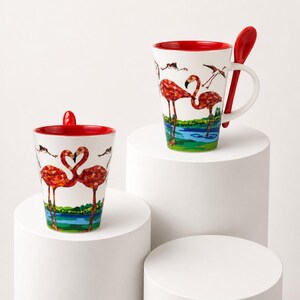 Peut inclure: Deux tasses en céramique blanche ornées d'un motif de flamants roses de style mosaïque. Les tasses ont un intérieur rouge vif et une forme conique. L'une des tasses comprend une cuillère rouge insérée dans la poignée. Le décor montre des flamants.