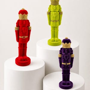 Puede incluir: Tres figuras de cascanueces con acabados de terciopelo en rojo vibrante, verde lima y morado intenso. Cada figura lleva una corona y presenta detalles de purpurina dorada en la corona, los puños y la hebilla del cinturón, sobre una base.