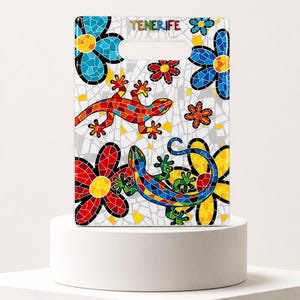 Op de afbeelding: Een rechthoekige snijplank met een kleurrijk mozaïekpatroon van twee hagedissen en diverse bloemen op een witte achtergrond. Het woord TENERIFE staat in zwarte letters gedrukt bij de handgreep aan de bovenkant. Het ontwerp bevat rode, blauwe, gele en groene tegels.