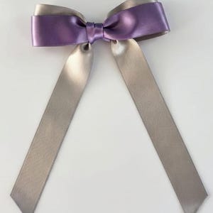 May include: A decorative bow crafted from smooth, lustrous satin ribbon. The design features a layered appearance with a vibrant purple ribbon positioned over a metallic silver ribbon, tied together in the centre with a matching purple knot.