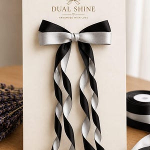 Puede incluir: Un lazo para el cabello de doble capa con una capa superior de satén negro y una capa inferior de satén plateado, sujeto a una tarjeta de presentación color crema con el texto DUAL SHINE y HANDMADE WITH LOVE, con dos cintas largas colgantes.