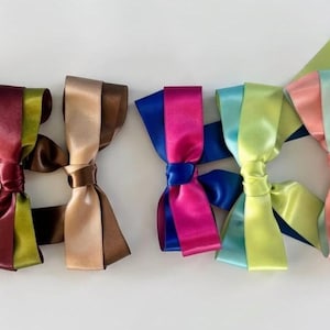 May include: A collection of six decorative satin ribbon bows arranged in a row. The bows feature various colour combinations including lavender, burgundy, olive green, brown, beige, magenta, royal blue, lime green, teal, and soft pink.