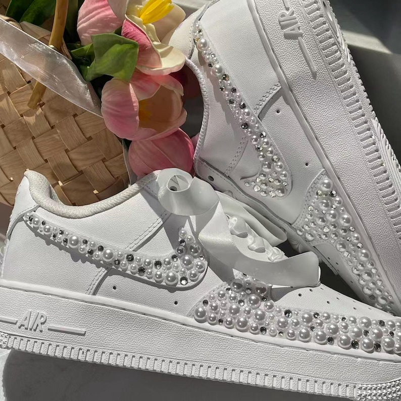 May include: A pair of white sneakers featuring wide satin ribbon laces and decorative embellishments consisting of small white pearls and clear crystals arranged along the side swoosh and the front toe box area of the footwear.