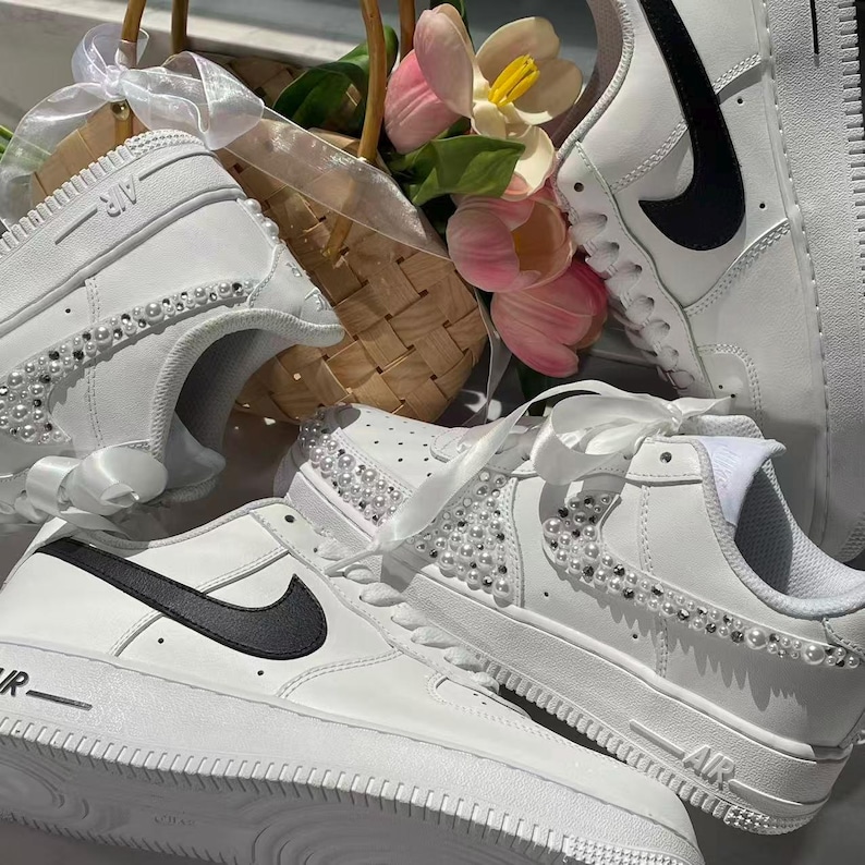 May include: White sneakers featuring black swoosh logos and decorative pearl embellishments along the sides. The shoes include white satin ribbon laces and the text AIR embossed on the soles. A woven basket with pink tulips is positioned behind the shoes.