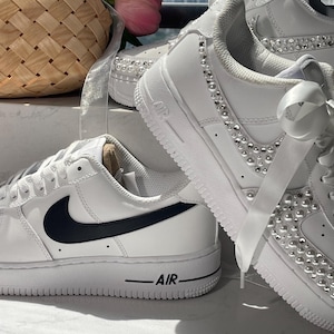 May include: A pair of white sneakers featuring a black Nike swoosh logo on the side and a thick white sole with the word AIR printed in black. Another white sneaker is decorated with silver studs and white pearls, featuring white ribbon laces.