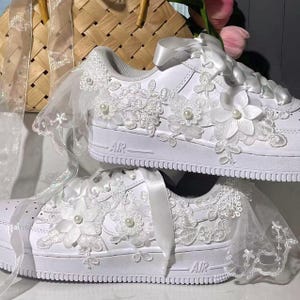 May include: A pair of white sneakers decorated with white floral lace appliques, small faux pearls, and sheer white ribbon laces. The shoes feature a delicate white tulle veil attached to the heel area, creating a layered and textured appearance.