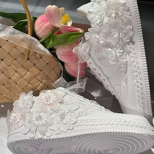 May include: A pair of white sneakers decorated with white lace floral appliques and trim, positioned near a woven basket containing pink tulips. The sneakers feature a textured rubber sole and intricate fabric flower details on the side panels.