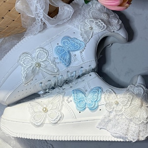 May include: A pair of white sneakers decorated with light blue and white embroidered butterfly appliques, white lace trim, and small pearl accents. The shoes feature white laces and a layered design with floral lace details on the heel and collar.
