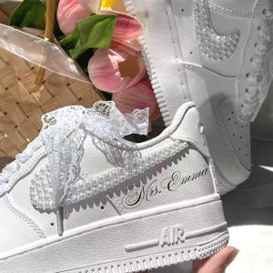 May include: A pair of white sneakers featuring white lace ribbons as shoelaces, pearl embellishments along the side swoosh and collar, and the text "Mrs. Emma" in black cursive script on the outer heel area of the shoe.